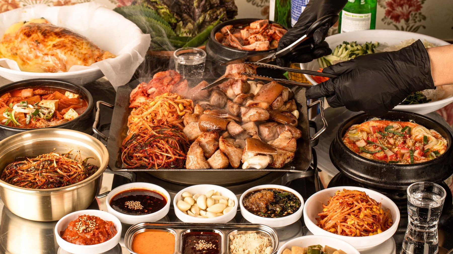 불판을 중심으로 반찬과 찌개가 둘러싼 한 상, 집게로 고기를 집는 장면